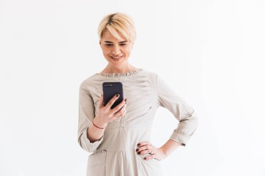 Elbise holding ve izole Studio beyaz arka plan üzerinde siyah smartphone kullanarak kısa sarı saçları zarif orta yaşlı kadınla