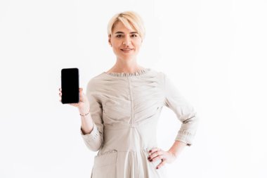 Studio beyaz arka plan üzerinde izole kamera elbise holding siyah smartphone ve gösteren boşaltmak ekran giyen kısa sarı saçları çekici orta yaşlı kadın