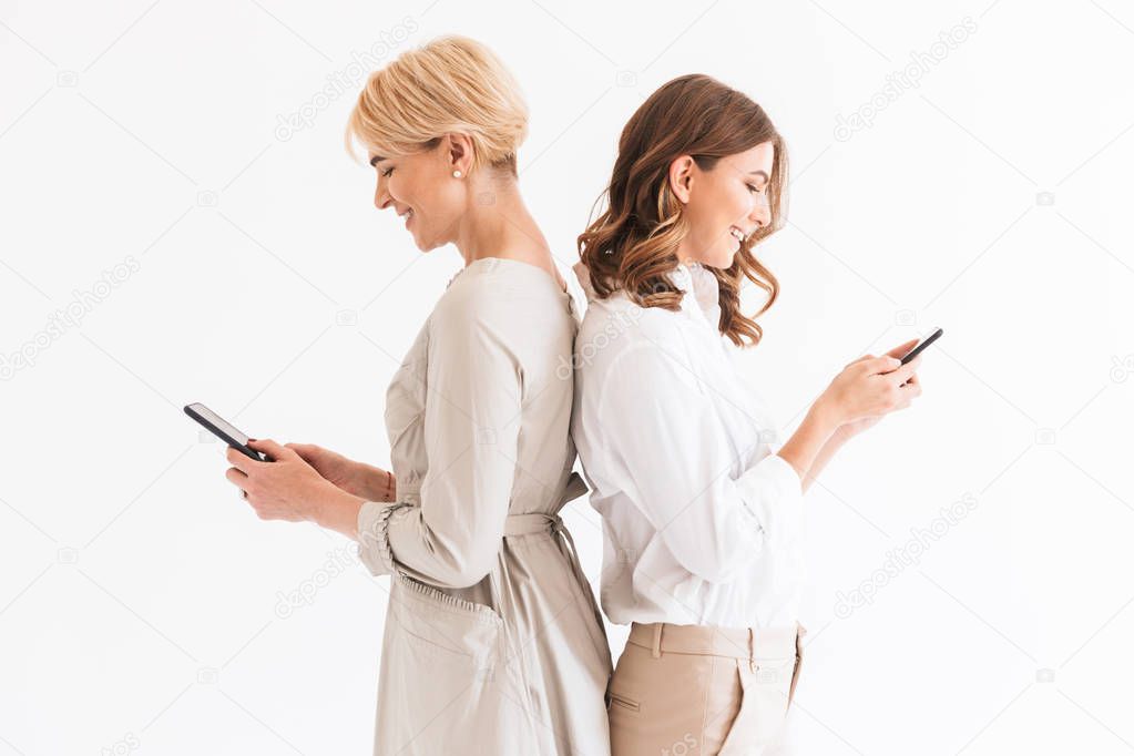Portrait of two happy women mother 40s and daughter 16-18 standing back to back and holding black smartphones together standing isolated over white background