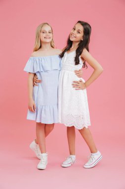 Elbiseler giymiş esmer ve sarışın kız tam uzunlukta fotoğraf gülümseyen ve sarılma birlikte pembe arka plan üzerinde izole ederken kameraya bakıyor