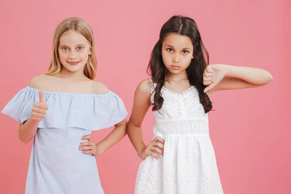 Like and dislike concept. Brunette and blonde girls 8-10 years old in dresses expressing different emotions with showing thumb up and down isolated over pink background