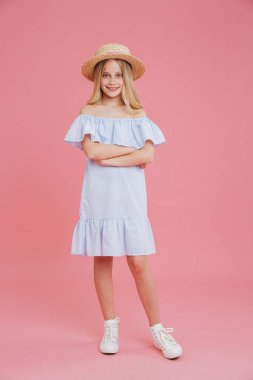 Tam uzunlukta portresi güzel sarışın yaz kız 8-10 giyiyor mavi elbise ve silah ile kamera poz hasır şapka geçti ve pembe arka plan üzerinde izole gülümse