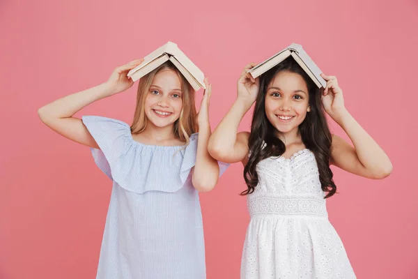 stock image Picture of happy european girls wearing dresses holding books at their heads while smiling at camera isolated over pink background