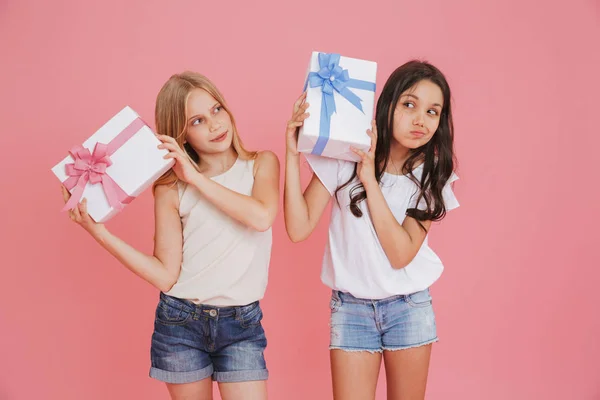 Two curious interested girls 8-10 in casual clothing holding and shaking gift boxes with colorful bows isolated over pink background