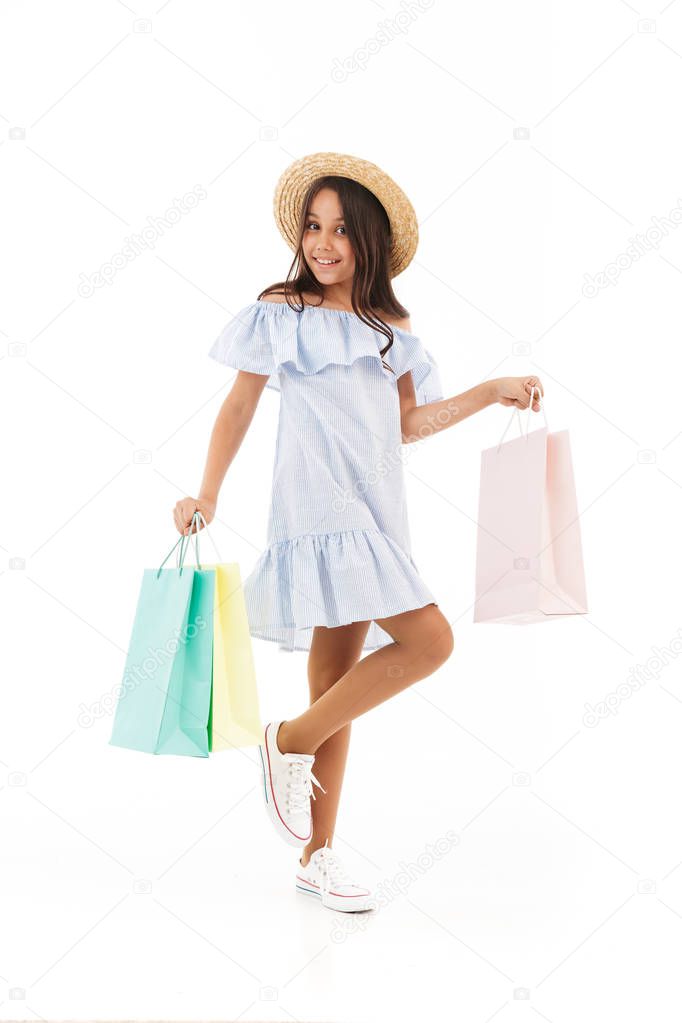 Full length image of Smiling young brunette girl in dress and straw hat posing with packages and looking at the camera over white background