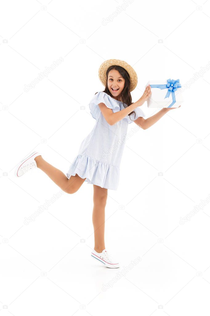 Full length image of Cheerful young brunette girl in dress and straw hat holding gift box while having fun and looking at the camera over white background