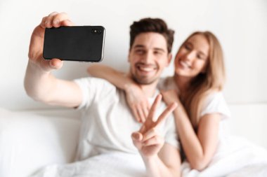 Yalanların genç neşeli sevgi dolu çift yatakta görüntü almak bir yana barış hareketi gösterilen smartphone bakarak bir selfie.