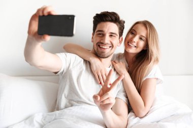 Yalanların genç neşeli sevgi dolu çift yatakta görüntü almak bir yana barış hareketi gösterilen smartphone bakarak bir selfie.