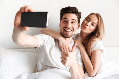 Neşeli sevgi dolu çift yalan yatakta bir kenara gösterilen smartphone bakarak bir selfie almak genç görüntü hareket başparmak.