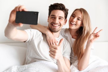 Yalanların genç neşeli sevgi dolu çift yatakta görüntü almak bir yana barış hareketi gösterilen smartphone bakarak bir selfie.