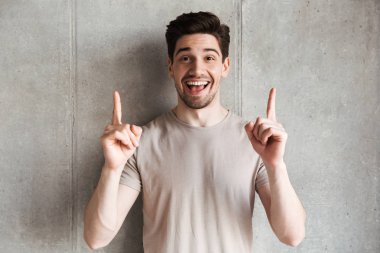 Mutlu genç adam gri duvar arka plan boşaltmak için işaret kamera seyir üzerinde ayakta fotoğrafı.
