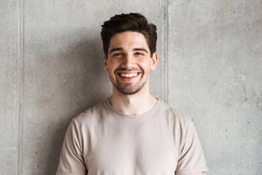 Image of happy young man standing over grey wall background looking camera.