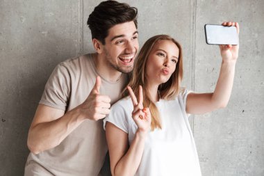 Görüntü iki neşeli insanlar adam ve kadın 20'li yaşlarda alarak selfie beton gri duvar üzerinde kapalı izole kamera el hareketi sırasında Smartphone cep telefonu ile fotoğraf