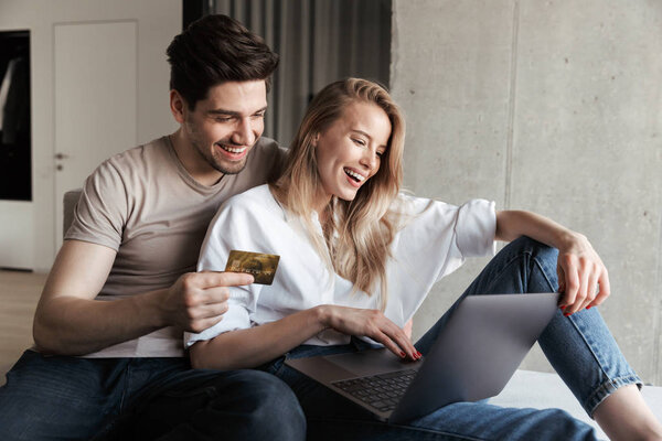 Photo of young happy loving couple in home indoors on sofa using laptop computer holding credit card.