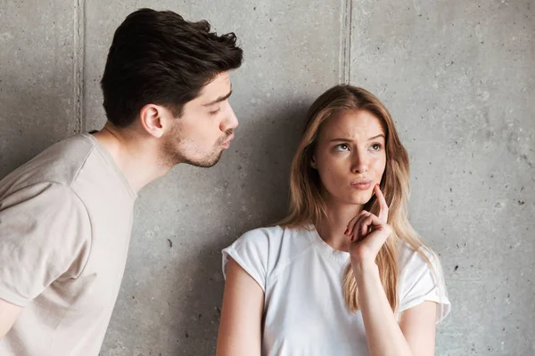 Photo of outraged doubtful woman hesitating while handsome man kissing her cheek isolated over gray background