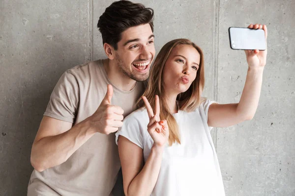 Image of two joyful people man and woman 20s taking selfie photo on smartphone while gesturing at camera isolated over concrete gray wall indoor