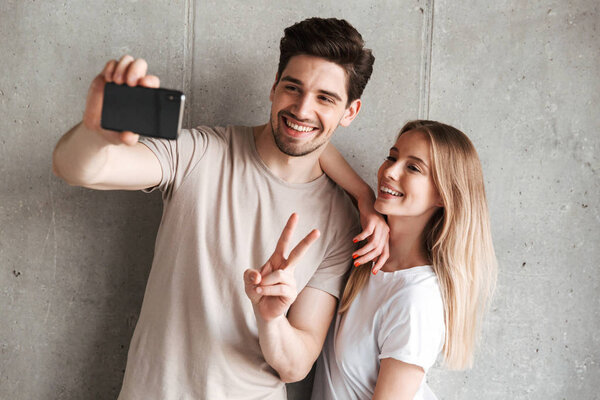 Image of two happy people man and woman taking selfie photo on smartphone while showing peace sign isolated over concrete gray wall indoor