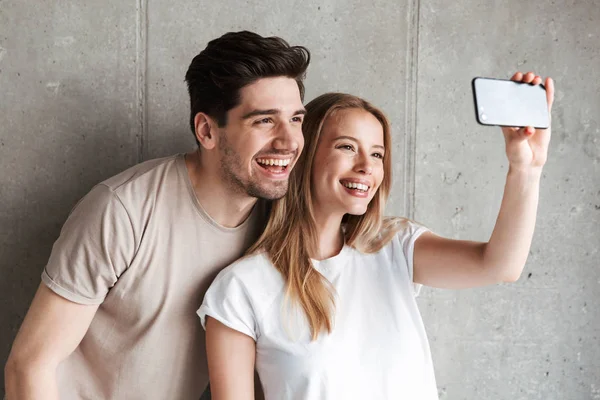 Image of two happy people man and woman taking selfie photo on smartphone while smiling at camera isolated over concrete gray wall indoor
