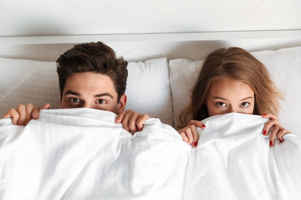 Image of young loving couple lies in bed covering faces with blanket looking camera.