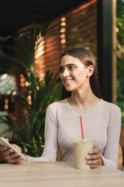 Bir kafede otururken açık havada bir smoothies ile cep telefonu kullanan genç kız gülümseyerek