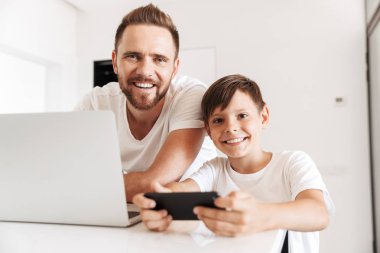 30'lu yaşlarda beyaz baba ve oğul 8-10 kameraya birlikte evde ise gümüş dizüstü kullanarak ve smartphone oyun gülümseyen portresi