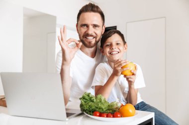 Mutlu fotoğrafı adam 30'lu ve çocuk mutfakta sebzeli yemek pişirme için 8-10 okuma tarifi laptop gülümseyen