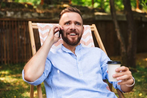 Portrait of handsome excited man 30s laughing and sitting in lounge chair with takeaway coffee during rest in park