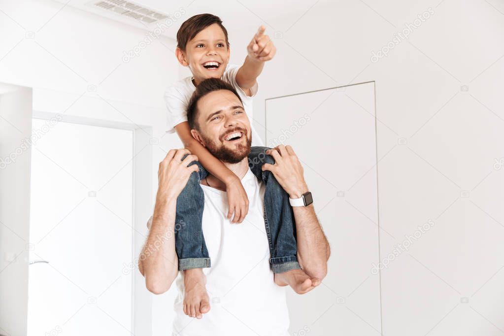 Photo of joyful happy boy 8-10 sitting on the neck of his father while looking aside indoor
