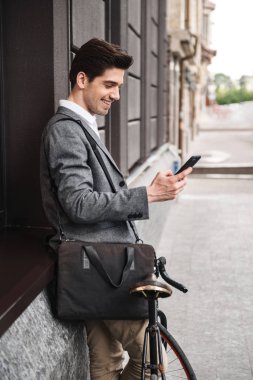 Genç işadamı açık havada bir bisiklet ile ayakta ve cep telefonuyla bir duvara yaslanmış gülümseyen