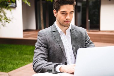 Açık havada otururken dizüstü bilgisayar üzerinde çalışan takım elbise giymiş ciddi genç işadamı