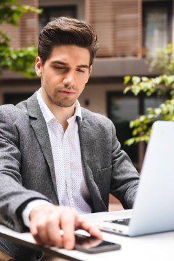 Ciddi genç işadamı cep telefonunuzun ve dizüstü bilgisayar açık havada oturan çalışırken takım elbise giymiş