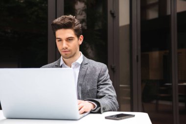 Açık havada otururken dizüstü bilgisayar üzerinde çalışan takım odaklı genç işadamı giymiş