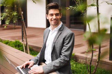 Açık havada otururken dizüstü bilgisayar üzerinde çalışan takım elbise giymiş neşeli genç işadamı