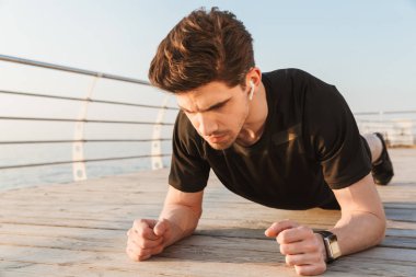 Tahta açık havada kulaklık ile müzik dinleme sahilde egzersiz spor yapmak imge-in konsantre yakışıklı genç sporcu.