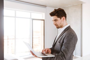 Evde ayakta iken dizüstü bilgisayar kullanan takım elbiseli ciddi genç adam