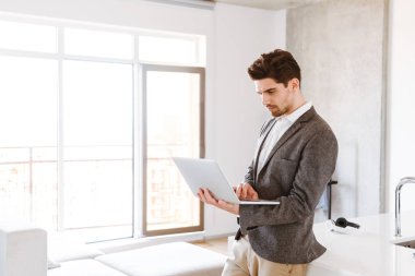 Evde ayakta iken dizüstü bilgisayar kullanan takım elbiseli ciddi genç adam