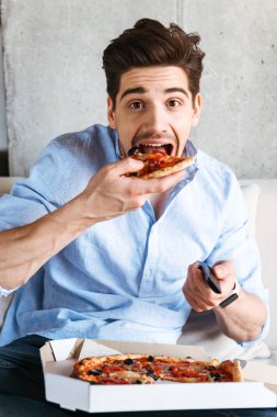 Evde bir kanepede oturan ve pizza yemek TV uzak tutan heyecanlı genç adam