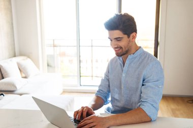 Evde otururken dizüstü bilgisayar kullanan genç gülümseyen bir adam portresi