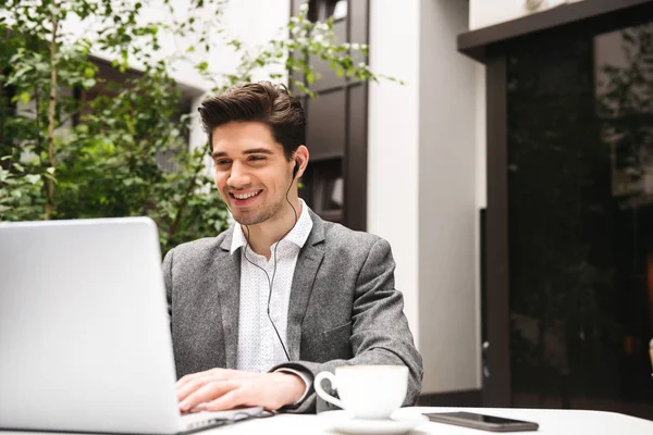 Portrait of a smiling young businessman in earphones looking at laptop computer while drinking coffee at the cafe outdoors