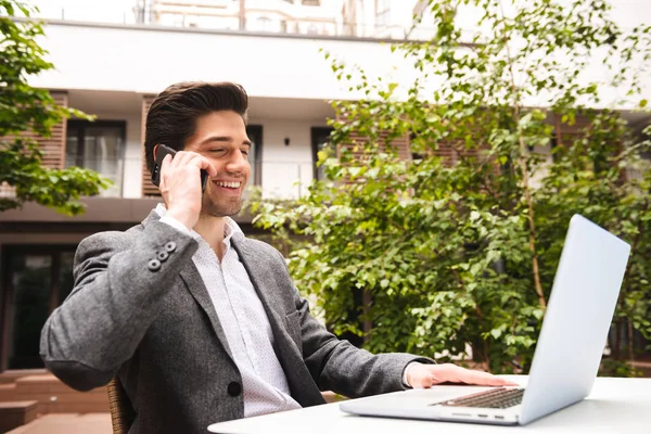 屋外に座ってラップトップ コンピューターで作業しながら携帯電話で話しているスーツに身を包んだ青年実業家を笑顔 — ストック写真