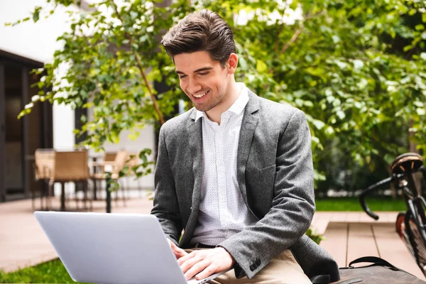 屋外に座ってラップトップ コンピューターで作業してスーツを着て笑っている青年実業家 — ストック写真