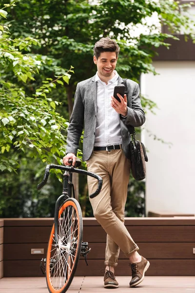自転車を屋外に立っている間携帯電話を使用してスーツを着て笑顔の青年実業家 — ストック写真