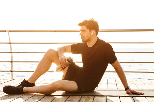 イヤホンで音楽を聴くビーチの上に水のボトルを屋外に座っている若いスポーツマンの写真 — ストック写真