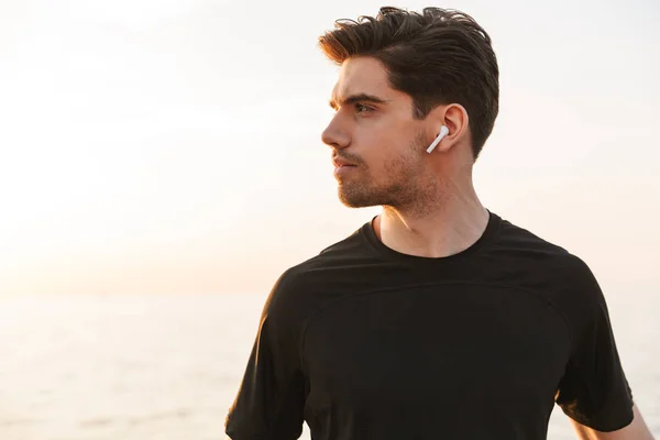 Charming young sportsman in black shirt in earphones standing at the seaside