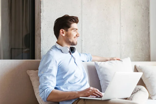 Joven Sonriente Con Auriculares Usando Portátil Mientras Está Sentado Sofá — Foto de Stock