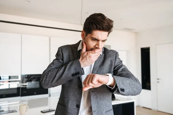 Shocked young man in suit looking at his wrist watch and covers mouth with hand at home