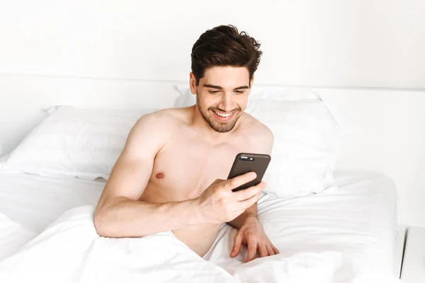 Smiling shirtless man using mobile phone while laying in bed at home