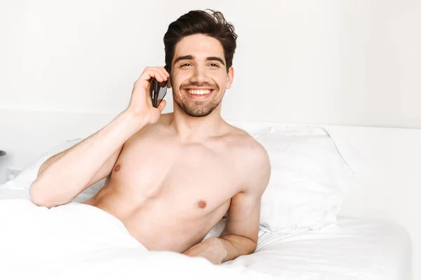 Smiling shirtless man talking on mobile phone while laying in bed at home