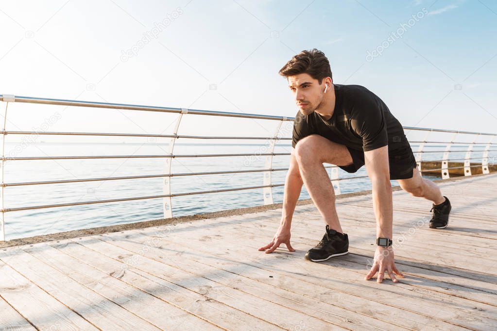 Photo of young concentrated sportsman outdoors on the beach listening music ready to run.