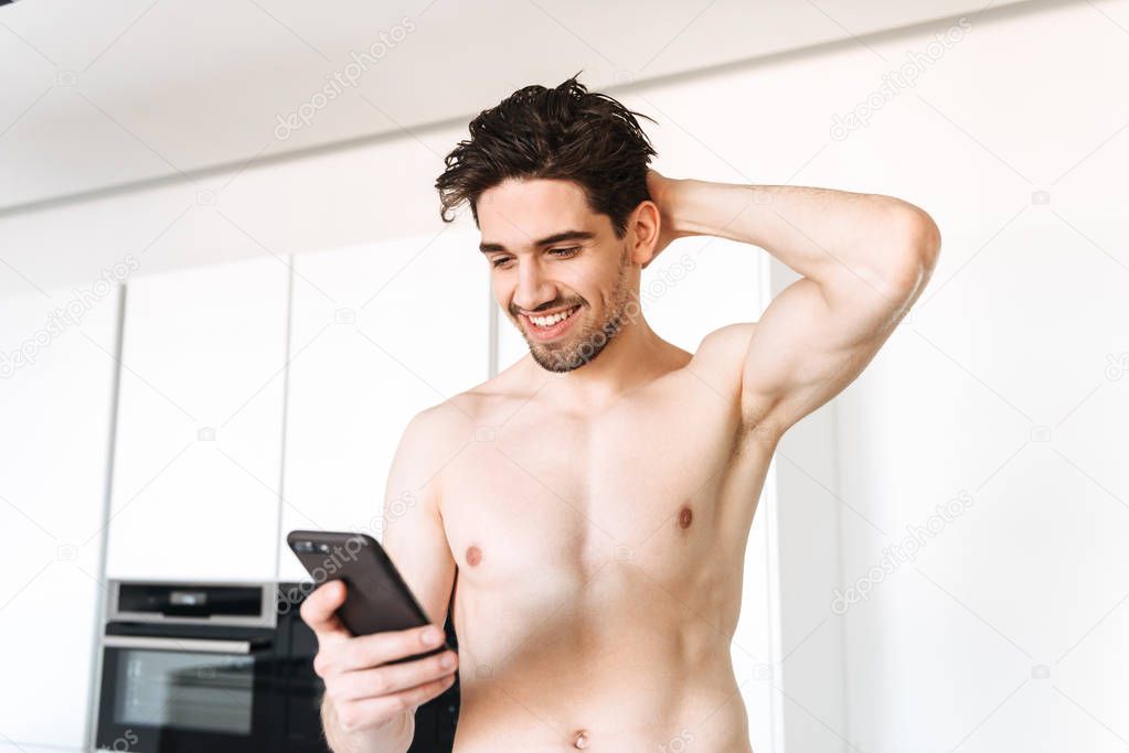 Photo of young cheerful naked man standing indoors at kitchen with towel. Looking aside using mobile phone.